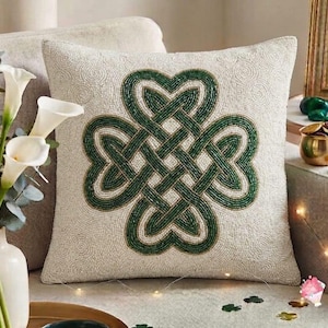May include: A square throw pillow with a cream-colored base featuring a central green beaded Celtic knot design. The entire surface of the pillow is covered in intricate, small beads arranged in swirling patterns, creating a textured appearance.