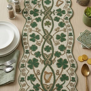 Può includere: Un runner da tavola allungato con bordi smerlati, caratterizzato da un'intricata lavorazione a perline nei toni del crema, verde smeraldo e oro. Il design mostra motivi ripetuti di trifogli, arpe dorate e piccoli cilindri, uniti da linee.