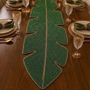 Peut inclure: Un grand chemin de table allongé en forme de feuille de palmier tropical, composé de nombreuses petites perles vert foncé. Le chemin de table présente une nervure centrale et des bords soulignés par une bordure de perles dorées contrastantes.