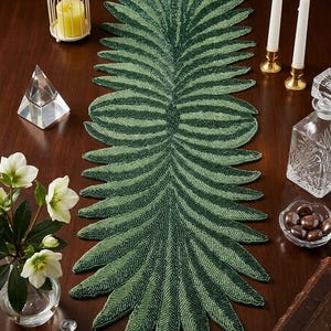 Op de afbeelding: Een groene, met kralen bezette tafelloper in de vorm van een blad. De loper heeft een donkergroene basis met lichtere groene accenten. Hij ligt op een donkerbruine houten tafel, samen met kaarsen en andere decoratieve items.