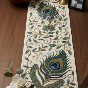 Puede incluir: Camino de mesa de cuentas color crema con diseño de plumas de pavo real. El camino de mesa presenta detalles en verde azulado, azul y dorado, con un patrón floral y de vid. Se muestra sobre una superficie de madera oscura.