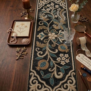 Pode incluir: Caminho de mesa preto com contas com um design floral e de pássaros em ouro, verde-azulado e branco. O caminho de mesa é rodeado por objetos decorativos, incluindo uma vela, uma bandeja com uma imagem emoldurada e um copo de água.