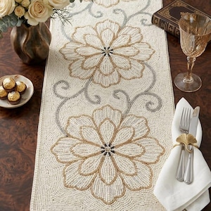 May include: Cream-colored beaded table runner with large floral designs in gold and gray. The runner features a repeating pattern of stylized flowers and scrolling vines, adding a touch of elegance to any dining table. The runner is approximately 14 inches wide.
