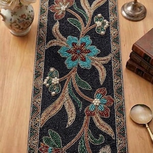 May include: A rectangular table runner featuring intricate beadwork on a dark background. The design displays a floral pattern with turquoise, red, and white beads, accented by pearl-like beads. A decorative border frames the central botanical motif.