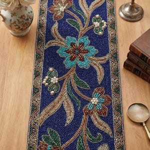 May include: A blue beaded table runner with a floral design. The runner features a central pattern of stylized flowers in teal, brown, and green, with pearl-like accents. The border is a detailed pattern of brown, green, and gold.