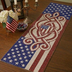 Puede incluir: Un camino de mesa patriótico con un diseño que presenta la bandera estadounidense y un escudo. El camino de mesa es principalmente azul, rojo y blanco, con una textura de cuentas. Una bandera estadounidense y flores están sobre la mesa.