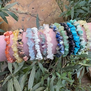 Puede incluir: Una colección de pulseras de virutas de piedras preciosas en varios colores, incluyendo naranja, amarillo, rosa, blanco, azul, verde y negro. Las pulseras se exhiben en un cilindro rosa con una banda dorada.