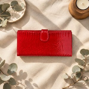 May include: A rectangular red wallet with a glossy, textured finish resembling reptile skin. The wallet features a central flap closure with a matching red snap button. The surface has a high-shine, patent-like appearance and a consistent, repeating pattern.