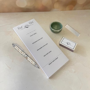 Puede incluir: Un bloc de notas blanco con las palabras "Reset Ritual" y preguntas para la reflexión, un bolígrafo con un diseño celestial, un cuenco verde pequeño, una varita de selenita y una caja de cerillas con un diseño de sol y luna.