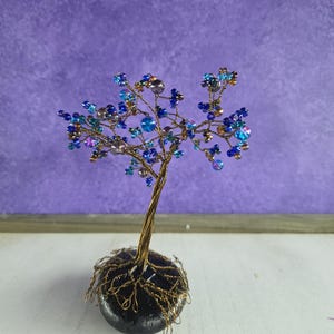 May include: A handcrafted wire tree sculpture with a gold-colored trunk and branches. The tree's leaves are made of blue, teal, and iridescent beads. The tree is rooted in a dark gray stone base, creating a unique decorative piece.