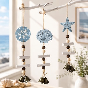 May include: Coastal-themed hanging decorations featuring a sand dollar, seashell, and starfish in light blue. Each ornament is suspended from a white branch with wooden beads, white sticks, and small brass bells. The beach-inspired decor adds a touch of the ocean to any space.