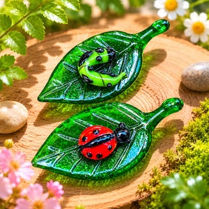 May include: Two glass leaf-shaped ornaments, each featuring a raised insect figure. One leaf holds a bright green caterpillar with black spots, while the other leaf displays a vibrant red ladybug with black spots and a black head.