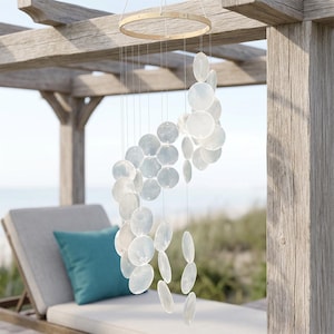 Peut inclure: Un carillon éolien circulaire composé de multiples disques de coquillages blancs, translucides et irisés, suspendus à un cerceau en bois clair par de fines cordes transparentes. Les disques sont disposés en spirale en cascade.