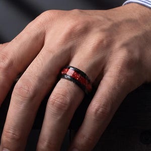 Op de afbeelding: Een zwarte ring met een centrale inleg van vermalen rood materiaal. De band heeft een gladde, donkere afwerking en een plat profiel, gedragen om een vinger. Het ontwerp bevat een levendig rood, getextureerd midden dat contrasteert met de zwarte randen.
