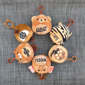 May include: A collection of plush keychains in various animal designs, including a bear, tiger, and elephant. Each keychain is a different color and has a metal clasp for attaching to keys or bags. Some keychains have text.