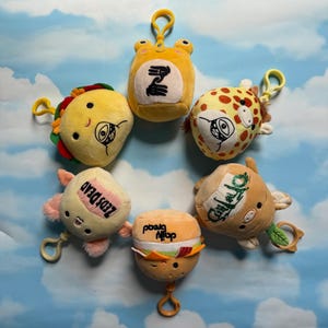 May include: A collection of plush toy keychains in various food and animal shapes, including a taco, frog, giraffe, and burger. Each keychain has a yellow clasp and cartoon-style facial features. The background is a blue sky with clouds.