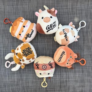 May include: A collection of plush keychains in various shapes and colors. The keychains include a cow with the text "GRIZ", a loaf of bread with the text "daily bread", and other cartoon characters. Each keychain has a metal clasp.