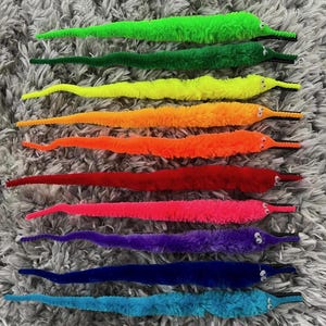 May include: A collection of colorful fuzzy worm toys in shades of green, yellow, orange, red, pink, purple, blue, and teal. Each worm has small white eyes and a black mouth. The toys are arranged on a gray, textured surface.