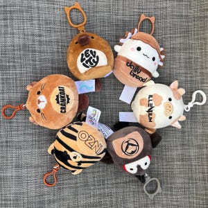 May include: A collection of plush keychains in various animal and food designs. The keychains feature brown, tan, and white colors, with text such as "CRANKDAT", "daily bread", and "OZNI". Each keychain has a clip for attachment.