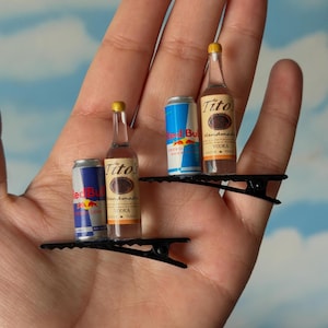 May include: Miniature Tito's Handmade Vodka bottles and Red Bull cans on black barrettes. The bottles have yellow caps and brown labels. The Red Bull cans are blue and silver. The barrettes are held in a hand.
