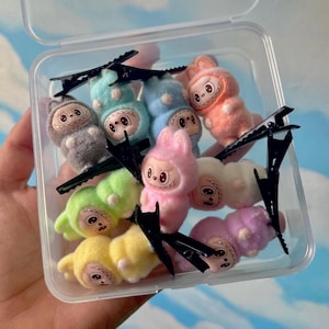 May include: A clear plastic container filled with colourful plush doll hair clips. The dolls are in various pastel colours, including pink, yellow, green, blue, and orange. Black metal hair clips are interspersed among the dolls.