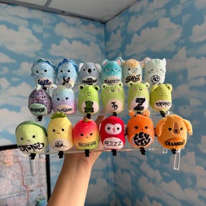 May include: A collection of twenty small, round plush toys arranged on a clear tiered display stand. Each plush features a unique character design, including animals, fruits, and abstract shapes, with various black text logos printed on their fronts.