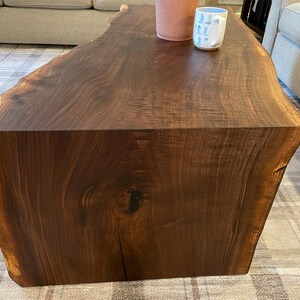 May include: A dark brown live edge wooden coffee table with a natural wood grain pattern, featuring a smooth surface. On the table sits a solid peach-colored cylindrical vase and a white ceramic mug decorated with a repeating blue cowboy boot pattern.