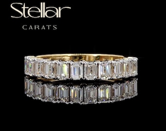 May include: A gold eternity band featuring a continuous row of emerald-cut clear stones set in a shared prong setting. The text Stellar CARATS is displayed above the ring, which is reflected on a dark surface, showcasing its metallic luster.