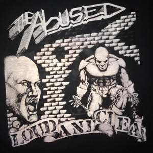 May include: Black t-shirt featuring a graphic design with the text "THE ABUSED" and "LOUD AND CLEAR". The design includes two bald figures, one screaming and the other chained, with a brick wall background.