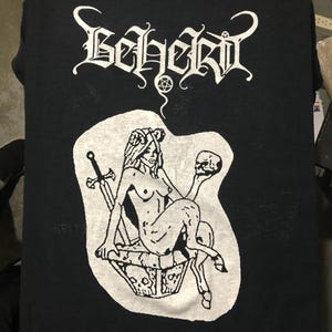 May include: Black t-shirt with a white graphic. The graphic features the word "Beherit" in a gothic font above an illustration of a nude figure holding a skull and a sword. The figure is seated on a decorated box.