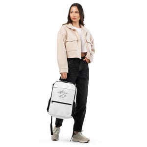 Könnte beinhalten: Ein weißer Rucksack mit einem schwarzen Bergmotiv und dem Text "Wander Often". Der Rucksack hat schwarze Träger und eine Vordertasche. Die Person trägt eine beige Jacke, ein weißes Oberteil und eine schwarze Jeans.