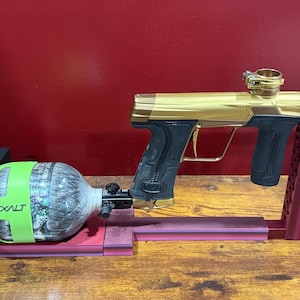 Può includere: Una pistola da paintball dorata e nera con un serbatoio d'aria verde e grigio. La pistola è montata su un supporto rosso e rosa. La parola "EXALT" è stampata sul serbatoio d'aria.