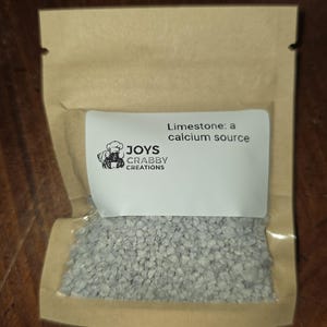 May include: A small, clear plastic bag filled with gray, granular limestone. The bag is sealed inside a brown paper pouch. A white label on the bag reads "Limestone: a calcium source" with the "JOYS CRABBY CREATIONS" logo.