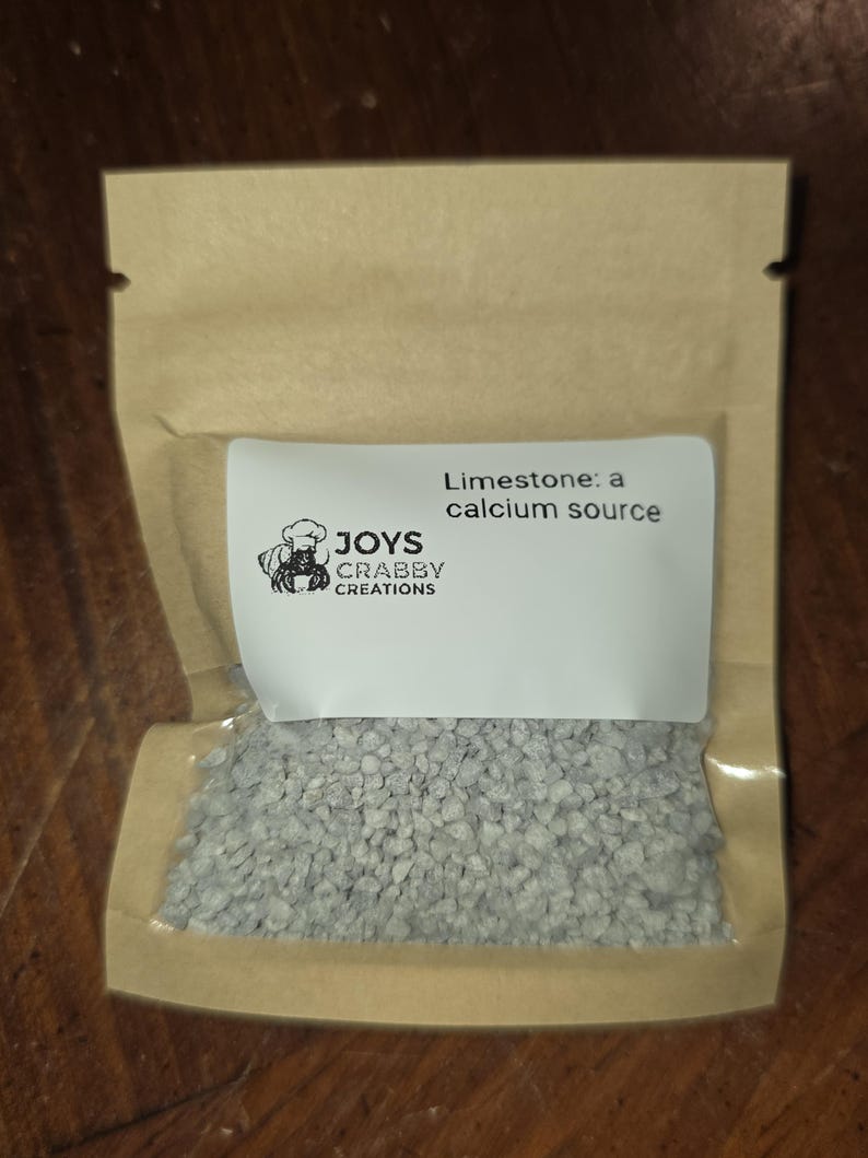 May include: A small, clear plastic bag filled with gray, granular limestone. The bag is sealed inside a brown paper pouch. A white label on the bag reads "Limestone: a calcium source" with the "JOYS CRABBY CREATIONS" logo.