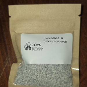 May include: A small, clear plastic bag filled with small, gray limestone rocks. A white label on the bag reads "Limestone: a calcium source" and features a crab logo with the text "JOYS CRABBY CREATIONS". The bag is sealed with a brown paper top.