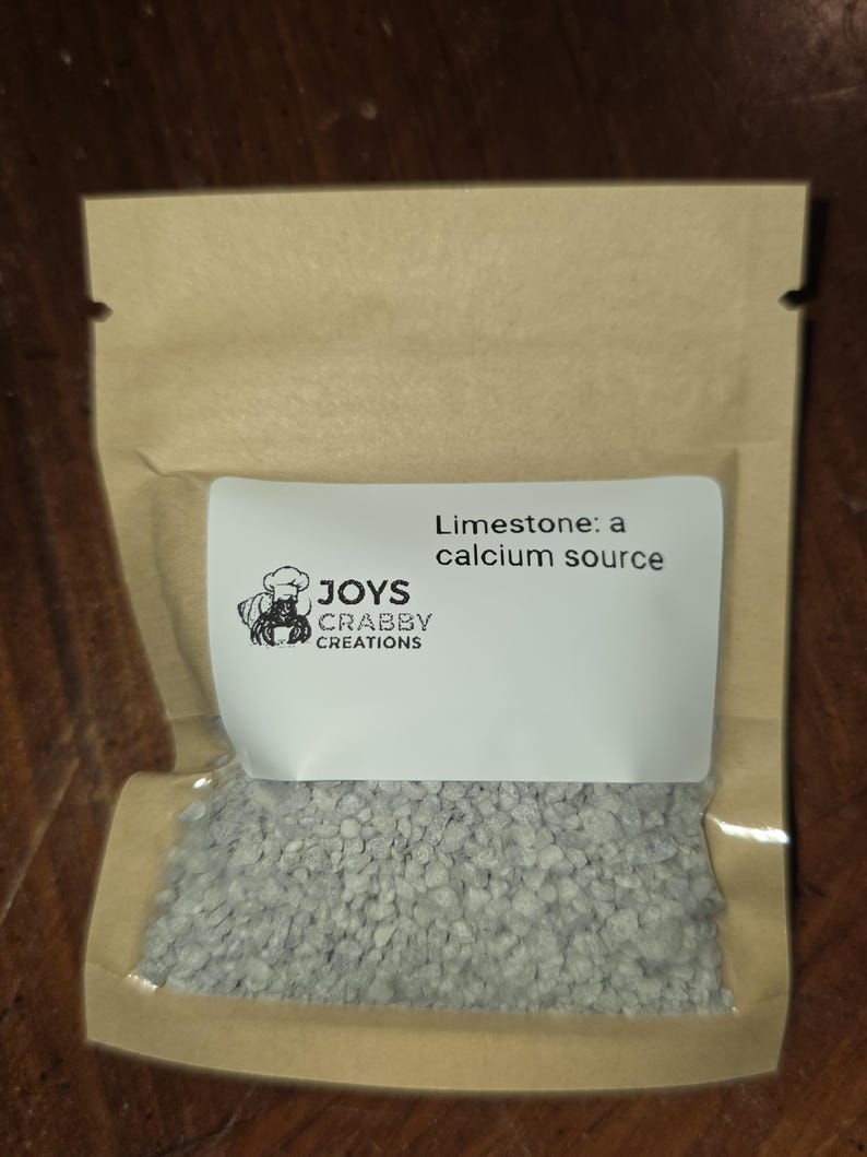 May include: A small, clear plastic bag filled with small, gray limestone rocks. A white label on the bag reads "Limestone: a calcium source" and features a crab logo with the text "JOYS CRABBY CREATIONS". The bag is sealed with a brown paper top.