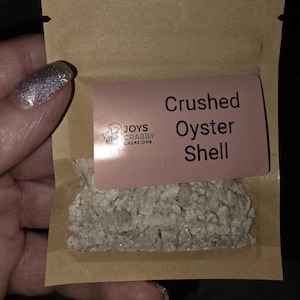 May include: A small brown paper bag containing crushed oyster shell. A pink label with the text "Crushed Oyster Shell" and the "Joys Crabby Creations" logo is affixed to the bag.