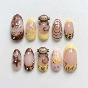 Puede incluir: Un conjunto de diez uñas artificiales con varios diseños. Las uñas presentan una mezcla de colores, incluyendo marrón, amarillo y rosa, con elementos decorativos como flores, espirales, estrellas y una figura de mono. Algunas uñas tienen pedrería.