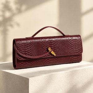 Könnte beinhalten: Eine rechteckige, tiefburgunderrote Clutch mit einer Schlangenleder-Prägung. Die Tasche verfügt über einen einzelnen Tragegriff, eine Umschlagklappe und ein goldfarbenes Metallknoten-Detail mittig auf dem Verschluss.