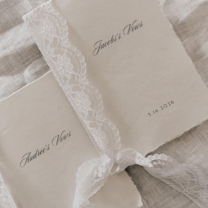 May include: Two cream-colored vow books tied with a white lace ribbon. The text "Jacob's Vows" and "Audree's Vows" are written in elegant script. The date "5.16.2016" is also visible. The books have a rustic, handmade appearance.