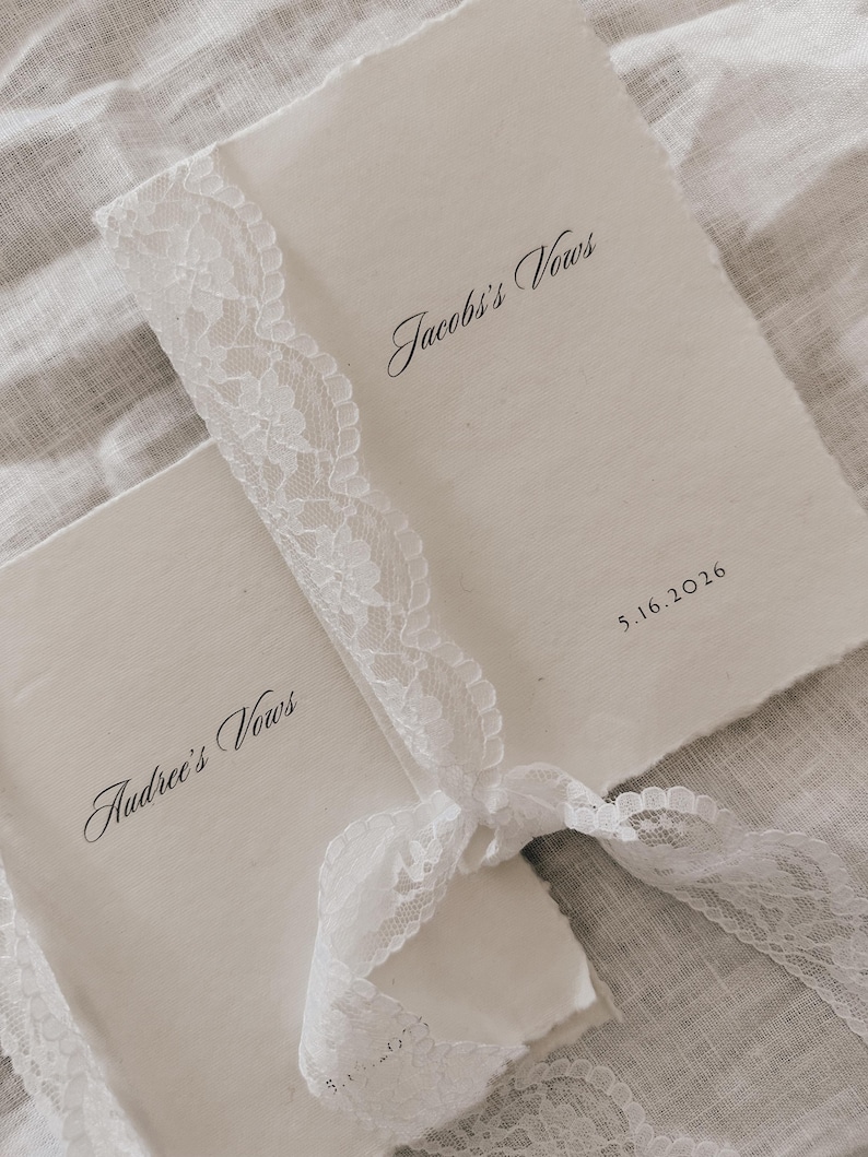 May include: Two cream-colored vow books tied with a white lace ribbon. The text "Jacob's Vows" and "Audree's Vows" are written in elegant script. The date "5.16.2016" is also visible. The books have a rustic, handmade appearance.