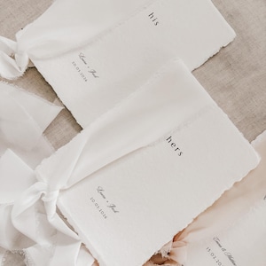 May include: White paper wedding cards with the words "his" and "hers" printed in black. Each card is tied with a white ribbon. The cards also include names and dates.