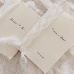 May include: Two off-white vow books with deckled edges, tied with a white lace ribbon. The text "Audree's Vows" and "Jacob's Vows" are printed in elegant script. The date "5.16.2026" is also printed on one of the books. The books rest on a textured, off-white surface.