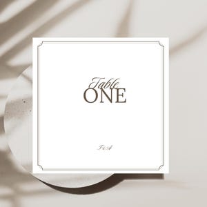 Op de afbeelding: Een witte tafelkaart met de woorden "Table ONE" in bruine script- en blokletters. De kaart heeft een dunne, decoratieve rand en rust op een lichtgekleurd, rond oppervlak. De schaduw van een plant wordt over de scène geworpen.