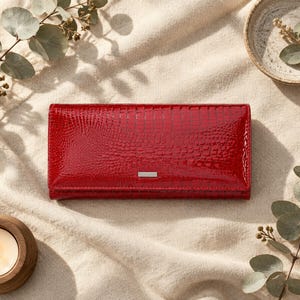 Puede incluir: Una cartera rectangular con un acabado rojo brillante y una textura de piel de cocodrilo. La cartera presenta una barra metálica horizontal delgada de color plateado centrada en la solapa frontal, aportando un detalle pulido.
