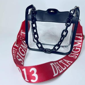 May include: Clear handbag with a black top and chain strap. The red shoulder strap features white text that reads "DELTA SIGMA 13". The bag has a clear body and a black chain strap.