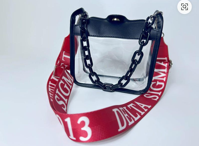May include: Clear handbag with a black top and chain strap. The red shoulder strap features white text that reads "DELTA SIGMA 13". The bag has a clear body and a black chain strap.