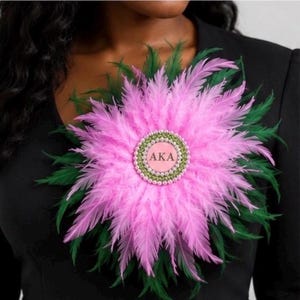 May include: A circular brooch featuring layers of vibrant pink and dark green feathers arranged in a sunburst pattern. At the center is a round pink disc with the letters AKA printed in black, surrounded by a border of small, sparkling green crystals.