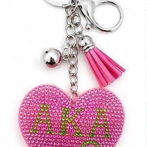 May include: A heart-shaped keychain featuring a pink rhinestone-covered surface with the letters A.K.A. spelled out in green rhinestones. The charm hangs from a silver-toned metal ring and chain, accompanied by a small silver ball and a pink tassel.