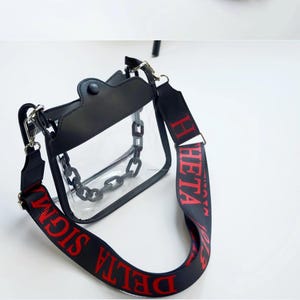 May include: A clear, rectangular crossbody bag with a black top and chain detail. The black strap features red text that reads "DELTA SIGMA THETA". The bag has a black chain inside.